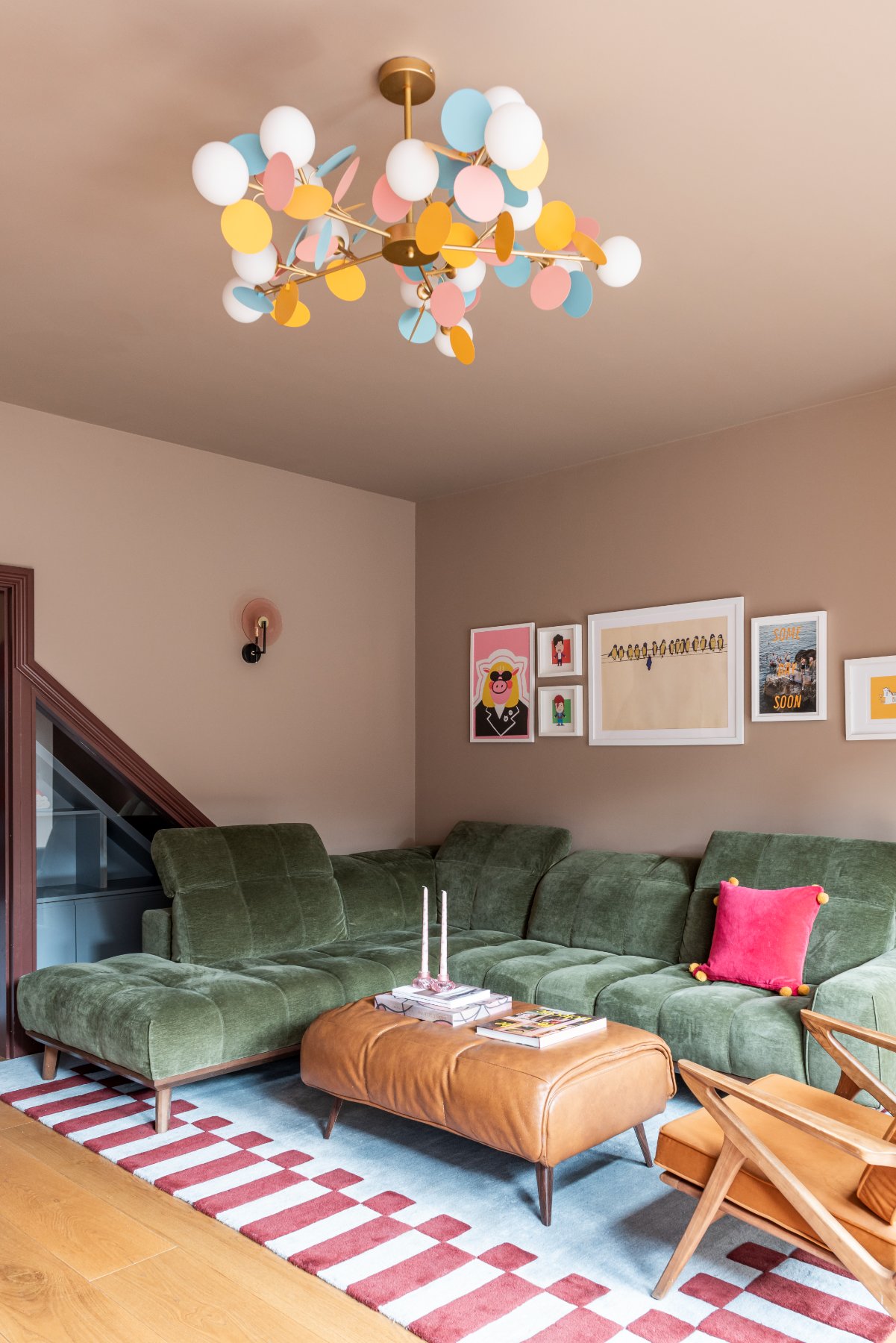Living room with a green velvet corner sofa, a tan leather ottoman, a striped rug and a colourful disc chandelier